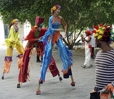 A group of performers wearing colorful costumes and face paint are walking on stilts in an outdoor setting. Trees and a historical building form the background, and one performer plays a trumpet while others interact joyfully.