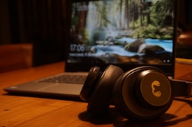 On a wooden table, a pair of black headphones rests in front of an open laptop. The laptop screen displays a nature-themed wallpaper with a forest and a stream.