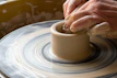 A close-up of a potter’s hands shaping a clay heart on the wheel, surrounded by bright glazes.