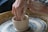 Hands are shaping a clay pot on a spinning pottery wheel. The hands are covered in wet clay, indicating that the person is actively molding the pot. The pot is in the early stages and is small and round. The background shows more clay residue, suggesting a typical pottery studio setting.
