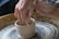 Hands are shaping a clay pot on a spinning pottery wheel. The hands are covered in wet clay, indicating that the person is actively molding the pot. The pot is in the early stages and is small and round. The background shows more clay residue, suggesting a typical pottery studio setting.
