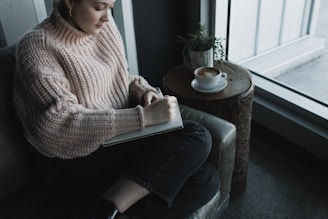 woman in white knit sweater and blue denim jeans sitting on gray sofa chair reading book