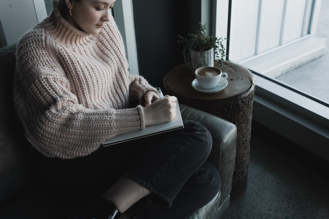 woman in white knit sweater and blue denim jeans sitting on gray sofa chair reading book