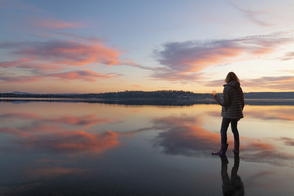 mulher na camisa branca da manga comprida em pé perto do lago durante o pôr do sol