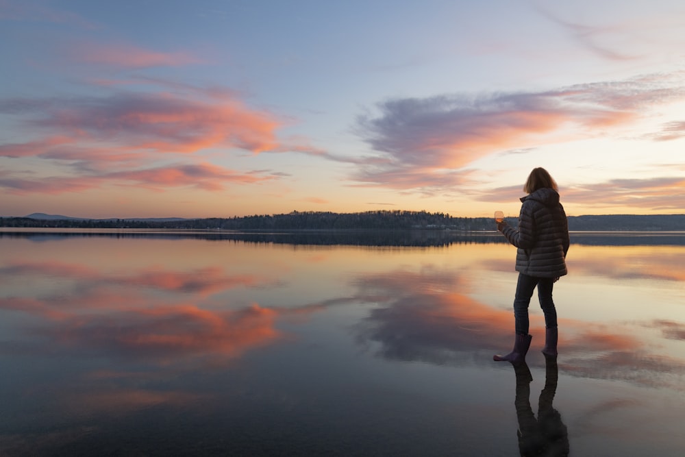 Mujer en camisa blanca de manga larga de pie cerca del lago durante la puesta del sol