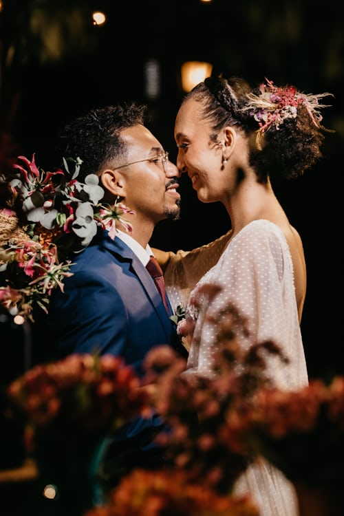 Un homme et une femme noire en tenue de mariage se regardent amoureusement. 
L'homme est plus petit que la femme, porte des lunettes, une barbe et des cheveux crépus. 
La femme est coiffée avec une tresse en couronne et une queue de cheval agrémentée de fleurs. Ses cheveux sont crépus également.