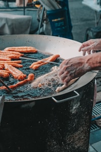 A large metal pan is filled with oil and several long dough sticks are being fried. Two hands are adjusting or placing the dough in the hot oil, surrounded by frying bubbles. The setting suggests an outdoor cooking or street food environment.