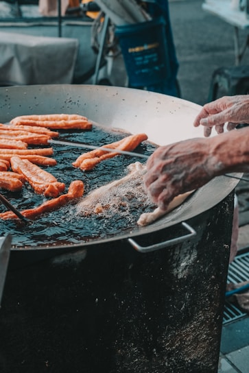 A large metal pan is filled with oil and several long dough sticks are being fried. Two hands are adjusting or placing the dough in the hot oil, surrounded by frying bubbles. The setting suggests an outdoor cooking or street food environment.