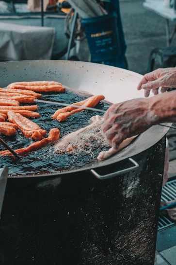 A large metal pan is filled with oil and several long dough sticks are being fried. Two hands are adjusting or placing the dough in the hot oil, surrounded by frying bubbles. The setting suggests an outdoor cooking or street food environment.