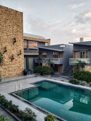 A modern residential complex with a large swimming pool in the foreground. The buildings are contemporary in design, featuring grey walls and wooden accents. The wall on the left side is adorned with artistic climbing sculptures. There is lush greenery around the pool area, including several potted plants and bushes.