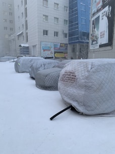 A snow-covered urban area with parked cars wrapped in protective covers. These cars are lined up along the side of a commercial building with various advertisements and posters on its wall. The environment appears to be cold, with snow on the ground and lingering fog.