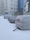 A snow-covered urban area with parked cars wrapped in protective covers. These cars are lined up along the side of a commercial building with various advertisements and posters on its wall. The environment appears to be cold, with snow on the ground and lingering fog.