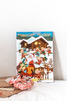 An advent calendar featuring a festive winter scene with Santa Claus and elves carrying gifts. Santa is in a sleigh surrounded by elves and reindeer, with a snowy log cabin in the background. The calendar is labeled with 'Calendrier de l'Avent Advent Calendar Chocolat au Lait Milk Chocolate'. In front of it, there are gift-wrapped presents with red and white patterned fabric.
