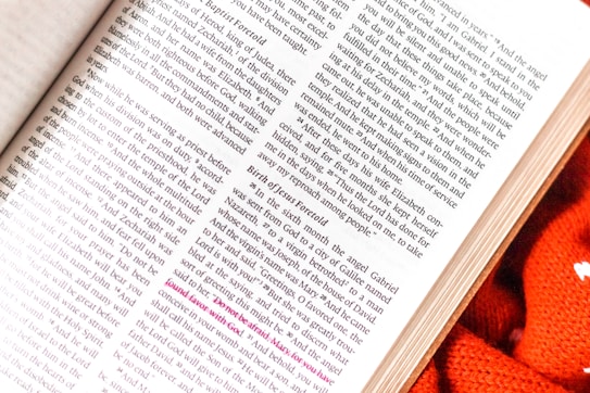 An open page of a book with text in a serif font occupying most of the space. The book appears to have religious or classical content, and some text lines are highlighted in pink. A section of the book is on a surface with a knitted orange fabric nearby.