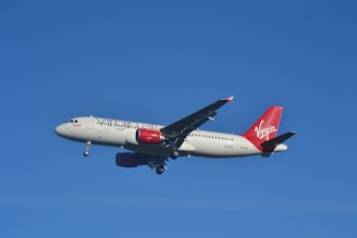 An airplane with distinctive red and white branding flies against a clear blue sky. The aircraft's landing gear is extended, indicating it is preparing for descent or landing.