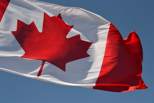 A large red maple leaf is centered on a white square background, with red vertical bars on either side. The flag is slightly rippling in the wind against a clear blue sky.