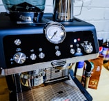 A close-up of a sleek, modern espresso machine with a shiny chrome finish. It has various dials and buttons for settings, and a pressure gauge prominently displayed on the front. Next to the machine, there is a metallic milk frothing pitcher and a package of coffee beans.