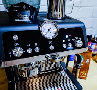 A close-up of a sleek, modern espresso machine with a shiny chrome finish. It has various dials and buttons for settings, and a pressure gauge prominently displayed on the front. Next to the machine, there is a metallic milk frothing pitcher and a package of coffee beans.