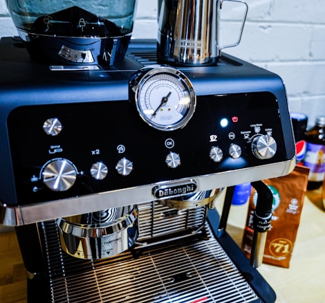 A close-up of a sleek, modern espresso machine with a shiny chrome finish. It has various dials and buttons for settings, and a pressure gauge prominently displayed on the front. Next to the machine, there is a metallic milk frothing pitcher and a package of coffee beans.