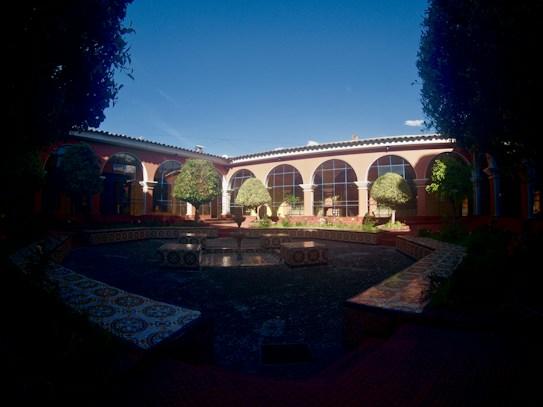 A courtyard featuring a central fountain surrounded by patterned tiles and bordered by neatly trimmed topiary. The building has large arched windows and is painted in warm colors, under a bright blue sky. Potted plants sit along the edges, enhancing the serene and elegant atmosphere.