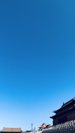 A clear blue sky stretches above traditional Chinese architecture. The rooftops of historical buildings are visible at the lower edge, characterized by their ornate eaves and wooden construction. In the distance, a modern skyscraper peeks above the skyline, contrasting with the ancient structures.