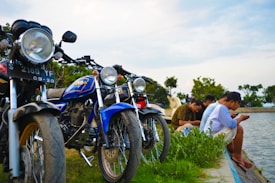 A group of motorcycles is parked on a grassy area by a body of water. Several people are seated near the water, engaged with their phones. The sky is partly cloudy, and trees can be seen in the background.