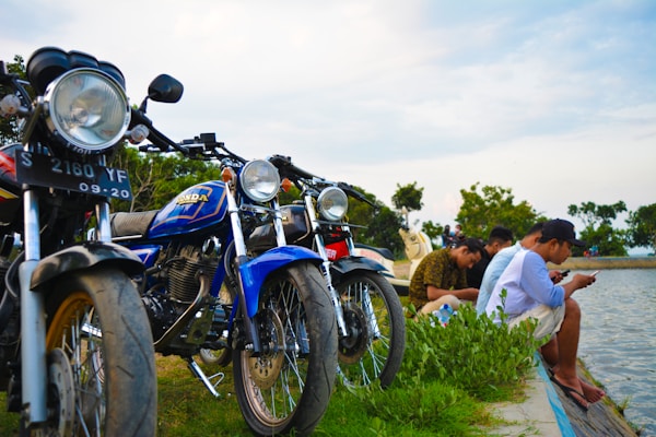 A group of motorcycles is parked on a grassy area by a body of water. Several people are seated near the water, engaged with their phones. The sky is partly cloudy, and trees can be seen in the background.
