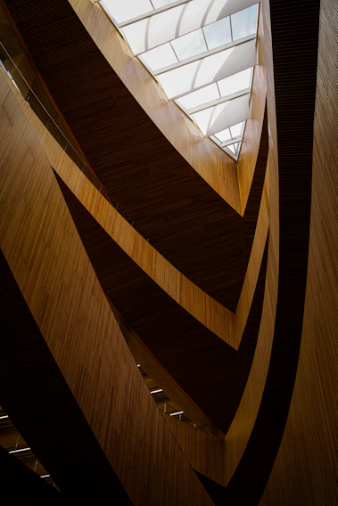 An architectural interior showcasing a modern, angular design with wooden slats that create a dynamic pattern. The ceiling features large, geometric skylights that allow natural light to stream in, adding contrast to the darker wooden tones.