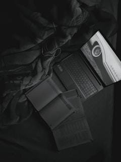 A grayscale image featuring a laptop displaying a screen with a circular object and numbers. Next to the laptop, open books and a notebook with a pen are visible. Items rest on a fabric surface that resembles a blanket.
