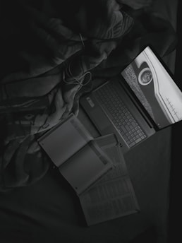 A grayscale image featuring a laptop displaying a screen with a circular object and numbers. Next to the laptop, open books and a notebook with a pen are visible. Items rest on a fabric surface that resembles a blanket.