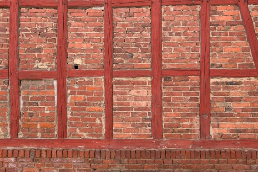 Brick wall with a rustic appearance featuring red wooden beams intersecting the brickwork in a traditional pattern. The bricks vary slightly in color, adding texture and depth.