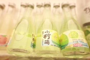 Several glass bottles with green labels are neatly arranged in a row. The labels feature Asian script and illustrations of green fruit, likely indicating a fruit-flavored beverage. The bottles have a soft, muted background, suggesting a focus on the middle row of bottles.