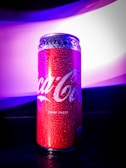 A red aluminum can with a glossy surface is covered in water droplets. The background features a gradient of purple and white light, creating a vibrant and modern aesthetic.