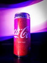A red aluminum can with a glossy surface is covered in water droplets. The background features a gradient of purple and white light, creating a vibrant and modern aesthetic.