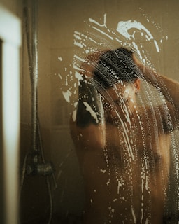 A person is seen through a glass shower door covered with soap suds. The individual is washing their hair, and the details are slightly obscured by the opaque soap patterns on the glass. The image has dim lighting, providing a private and intimate atmosphere.