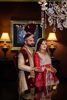 A groom in a deep maroon sherwani with gold accents, standing in a regal palace corridor.