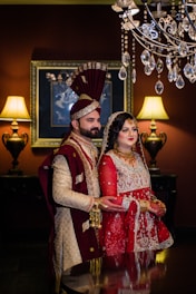 A couple dressed in traditional South Asian wedding attire stand in an elegantly decorated room. The man wears a cream and maroon sherwani with a matching turban, and the woman is dressed in a red and gold lehenga with intricate embroidery, adorned with jewelry. A chandelier with crystal accents hangs above them. Two ornate lamps on either side cast warm light, complementing the rich, dark tones of the room.