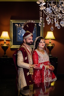 A couple dressed in traditional South Asian wedding attire stand in an elegantly decorated room. The man wears a cream and maroon sherwani with a matching turban, and the woman is dressed in a red and gold lehenga with intricate embroidery, adorned with jewelry. A chandelier with crystal accents hangs above them. Two ornate lamps on either side cast warm light, complementing the rich, dark tones of the room.