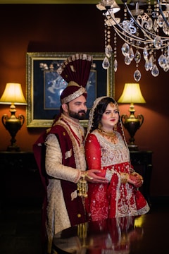 A couple dressed in traditional South Asian wedding attire stand in an elegantly decorated room. The man wears a cream and maroon sherwani with a matching turban, and the woman is dressed in a red and gold lehenga with intricate embroidery, adorned with jewelry. A chandelier with crystal accents hangs above them. Two ornate lamps on either side cast warm light, complementing the rich, dark tones of the room.
