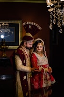 A couple in traditional South Asian wedding attire stands close together. The man wears an ornate sherwani with a matching turban, while the woman is dressed in an elaborately embroidered lehenga with intricate jewelry. Elegant lighting fixtures and a framed painting accentuate the background, creating a luxurious atmosphere.