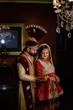 A couple in traditional South Asian wedding attire stands close together. The man wears an ornate sherwani with a matching turban, while the woman is dressed in an elaborately embroidered lehenga with intricate jewelry. Elegant lighting fixtures and a framed painting accentuate the background, creating a luxurious atmosphere.