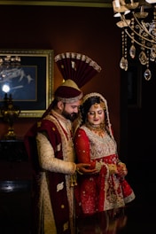 A couple in traditional South Asian wedding attire stands close together. The man wears an ornate sherwani with a matching turban, while the woman is dressed in an elaborately embroidered lehenga with intricate jewelry. Elegant lighting fixtures and a framed painting accentuate the background, creating a luxurious atmosphere.