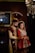 A couple in traditional South Asian wedding attire stands close together. The man wears an ornate sherwani with a matching turban, while the woman is dressed in an elaborately embroidered lehenga with intricate jewelry. Elegant lighting fixtures and a framed painting accentuate the background, creating a luxurious atmosphere.