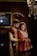 A couple in traditional South Asian wedding attire stands close together. The man wears an ornate sherwani with a matching turban, while the woman is dressed in an elaborately embroidered lehenga with intricate jewelry. Elegant lighting fixtures and a framed painting accentuate the background, creating a luxurious atmosphere.