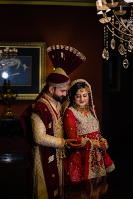A couple in traditional South Asian wedding attire stands close together. The man wears an ornate sherwani with a matching turban, while the woman is dressed in an elaborately embroidered lehenga with intricate jewelry. Elegant lighting fixtures and a framed painting accentuate the background, creating a luxurious atmosphere.
