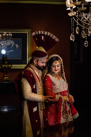 A couple in traditional South Asian wedding attire stands close together. The man wears an ornate sherwani with a matching turban, while the woman is dressed in an elaborately embroidered lehenga with intricate jewelry. Elegant lighting fixtures and a framed painting accentuate the background, creating a luxurious atmosphere.