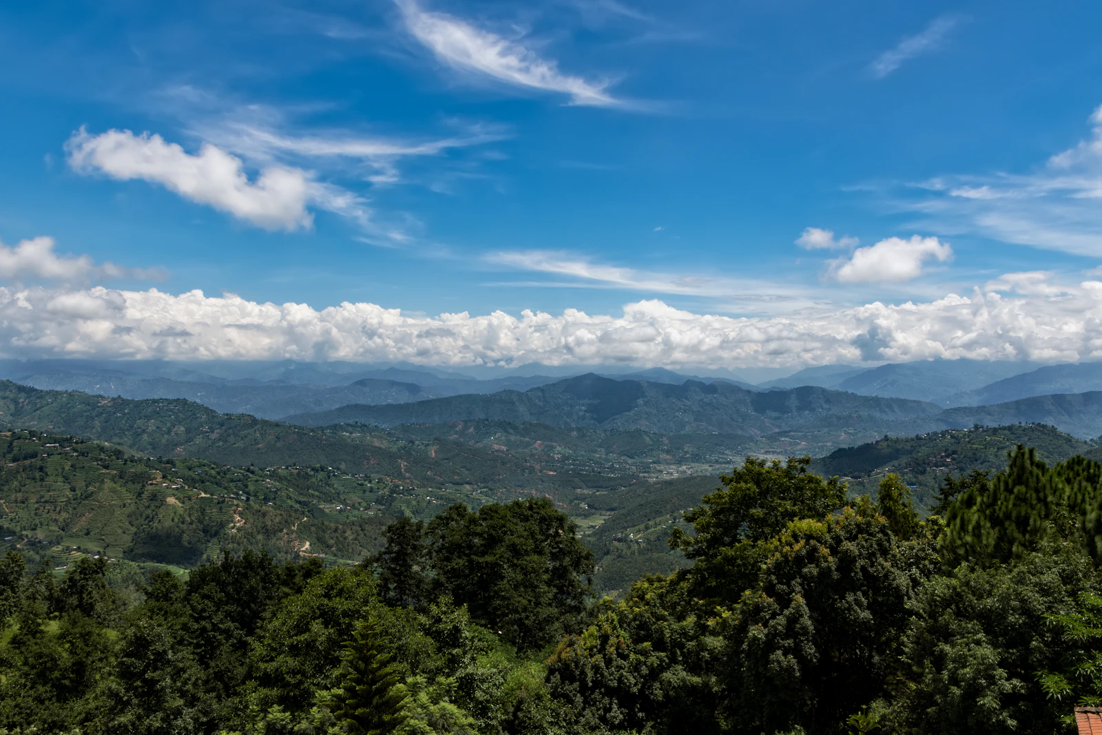 Forested ridges and terraced hillsides falling away from Dhulikhel, Nepal, under a bank of white cloud and deep blue sky