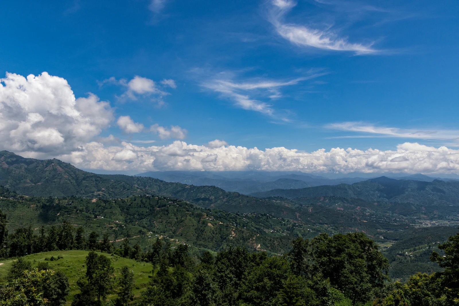 Forested ridges and terraced slopes falling away from Dhulikhel toward distant hills under a bright blue sky