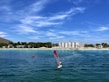 A windsurfer navigates the calm, clear blue waters near a bustling beach lined with trees and a large hotel. The sky is clear with some wispy clouds, complementing the vibrant summer atmosphere.