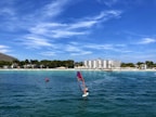 A windsurfer navigates the calm, clear blue waters near a bustling beach lined with trees and a large hotel. The sky is clear with some wispy clouds, complementing the vibrant summer atmosphere.